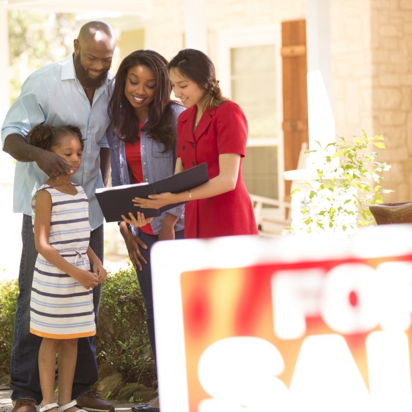 Latin descent realtor shows African descent family a new home to purchase.  Mother, father and daughter.  Real estate sign.  Home in background.  Spring or summer season.